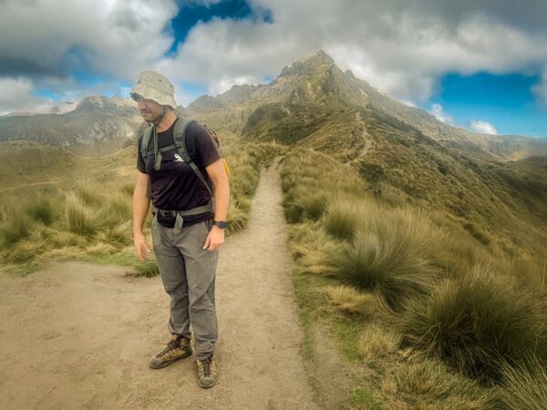 A hiker stands on a trail surrounded by lush green grass leading to Pichincha Volcano in Ecuador under a cloudy sky.