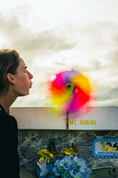 Colorful pinwheel spins while a woman blows on it at Tulcán Cemetery, Ecuador, with decorations and cloudy sky in the background.