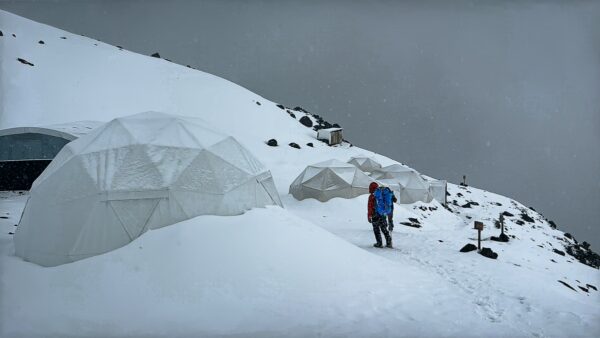 High camp domes blanketed in snow with a hiker in red near Chimborazo Volcano, Ecuador, against a cloudy gray sky.