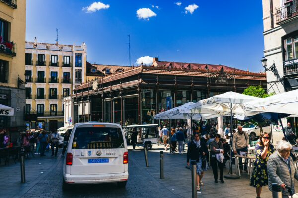 Bustling scene at Mercado de San Miguel, Madrid, with crowds, white market umbrellas, and historic architecture under a bright blue sky.