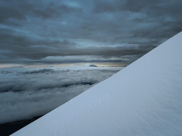 Snow-covered slopes of Mount Cotopaxi rise sharply against a dramatic sky with clouds and distant peaks visible in Ecuador.