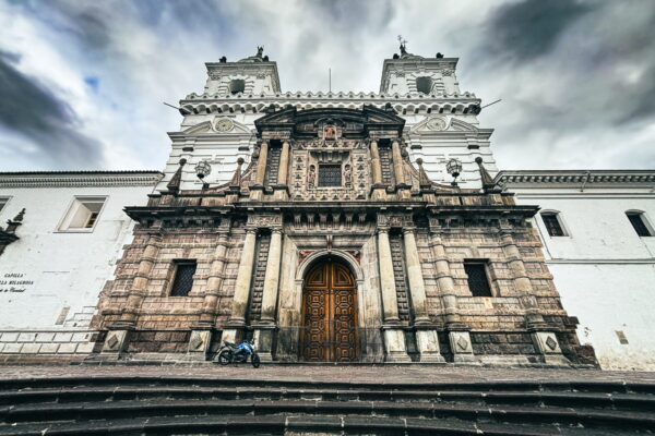 Intricate church façade with detailed stonework and wooden doors, set against a dramatic, cloudy sky in Quito, Ecuador.
