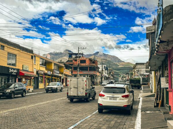 Bustling street scene in Machachi, Ecuador, featuring colorful buildings, parked cars, and distant mountains under a blue sky.