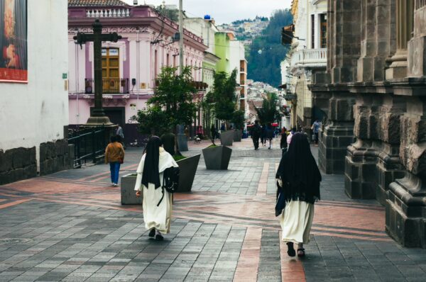 Nuns in traditional attire walk along a cobblestone street, flanked by colorful colonial buildings in Quito, Ecuador.