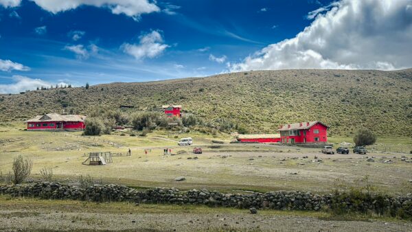 Bright red buildings stand against the lush, green landscape of Cotopaxi National Park, under a dramatic blue sky with scattered clouds.