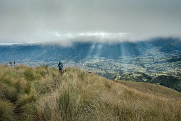Lush hills carpeted in grass descend towards the valley, with sunbeams breaking through clouds over Imbabura Volcano, Ecuador.