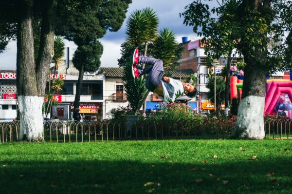A boy performs a backflip on the grass at Parque Ayora, surrounded by trees and colorful buildings in Tulcán, Ecuador.