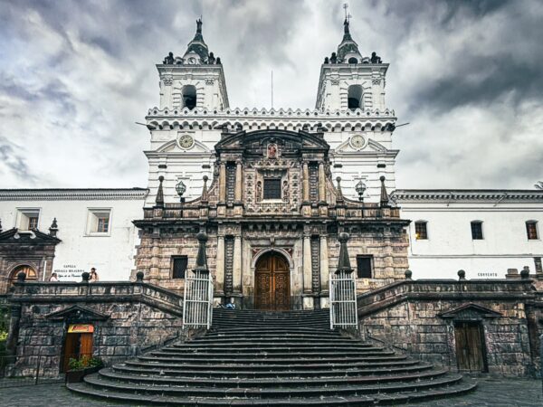 Elaborate stone church with ornate architecture and stairway leading to grand entrance, set against a cloudy sky in Quito, Ecuador.