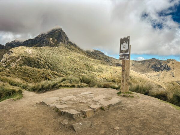 Pichincha Volcano features rugged terrain and a scenic viewpoint with a signpost against a backdrop of clouds and mountains in Ecuador.