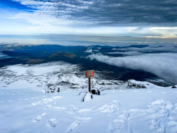 Snow-covered landscape with a sign at high camp on Chimborazo Volcano, offering panoramic views of clouds and distant mountains in Ecuador.