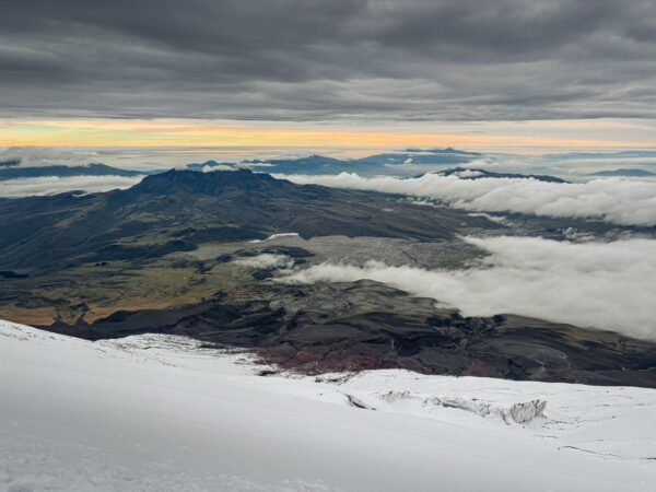Snow-capped Mount Cotopaxi towers above a landscape of rolling hills and clouds, bathed in soft morning light.
