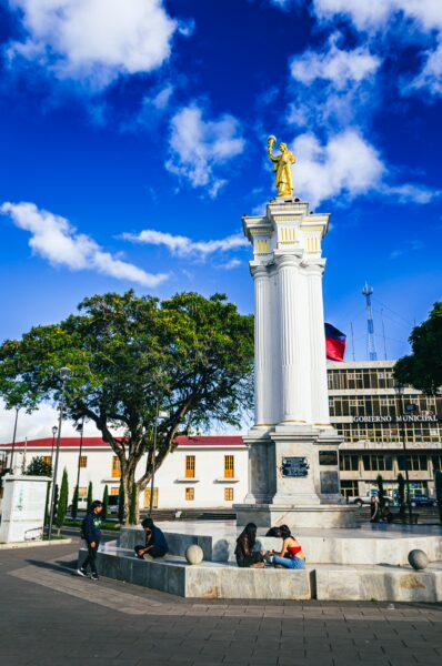 Central park in Tulcán features a white column topped by a golden statue against a vibrant blue sky and lush green trees.