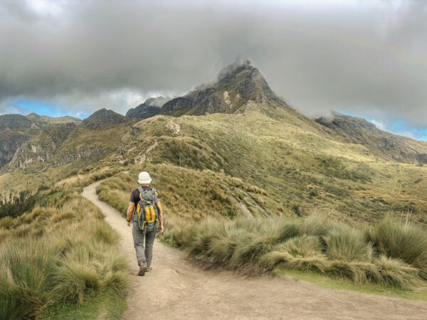 Hiker on a winding path through lush green grass, approaching the majestic Pichincha Volcano in Ecuador, shrouded in clouds.