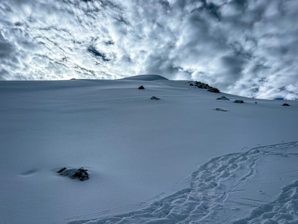 Snow-covered slopes of Chimborazo Volcano in Ecuador under a dramatic sky, featuring scattered rocks and footprints in the snow.
