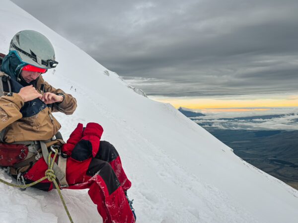 Climber in red gear adjusts equipment on the snowy slopes of Mount Cotopaxi, with a dramatic sky and distant landscape at sunset.