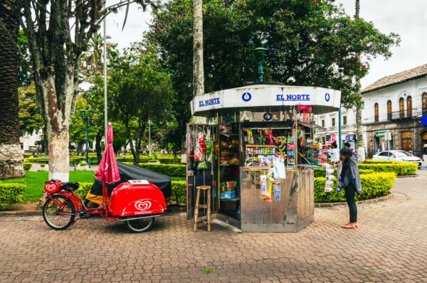 Colorful newsstand with vibrant magazines and snacks, situated in Parque Pedro Moncayo, Ibarra, Ecuador, surrounded by greenery.