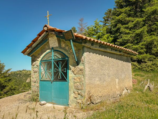 Small stone chapel with a green door and cross, surrounded by lush greenery in Lagadia, Arcadia, Greece. Bright blue sky above.