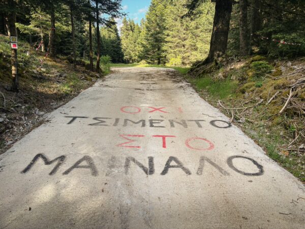 Graffiti reading "Όχι Τσιμέντο Στο Μαινάλι" on a forest road in Elati, Menalon, Arcadia, surrounded by lush greenery.