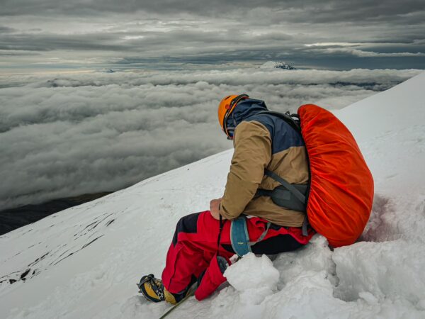 Climber in warm gear resting on snowy slopes of Mount Cotopaxi, surrounded by clouds and a dramatic gray sky.