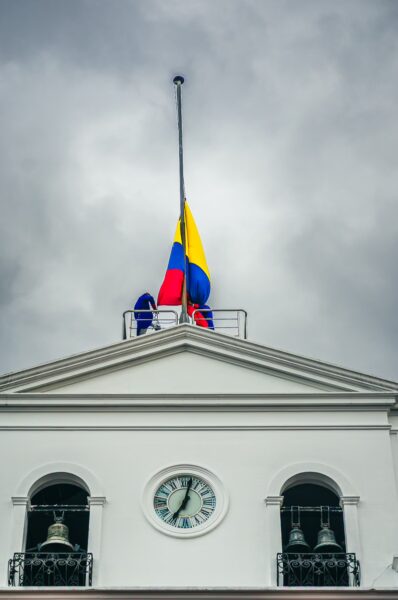 Carondelet Palace features a Colombian flag at half-mast under a cloudy sky, with a clock and decorative bell details visible.