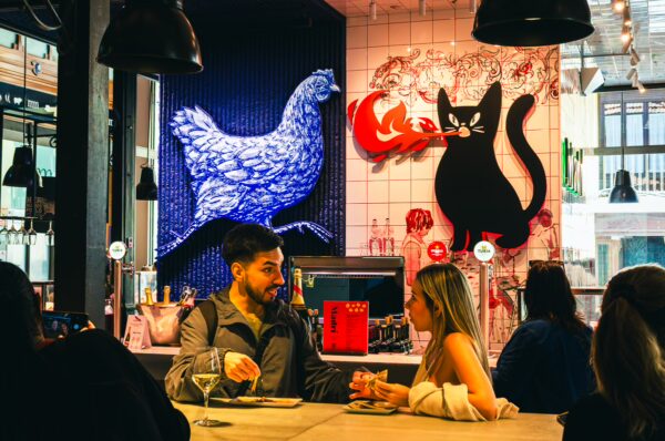Couple enjoying a meal at Mercado de San Miguel, Madrid, with vibrant artwork featuring a blue chicken and a black cat in the background.