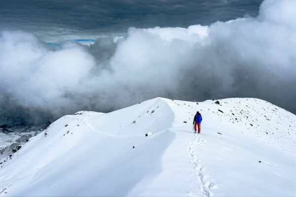 Snow-covered Chimborazo volcano in Ecuador with a lone hiker navigating fresh tracks under cloudy skies.