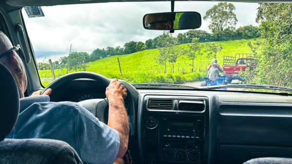 Driving through lush green fields in Machachi, Ecuador, with a rural road and a distant vehicle visible.