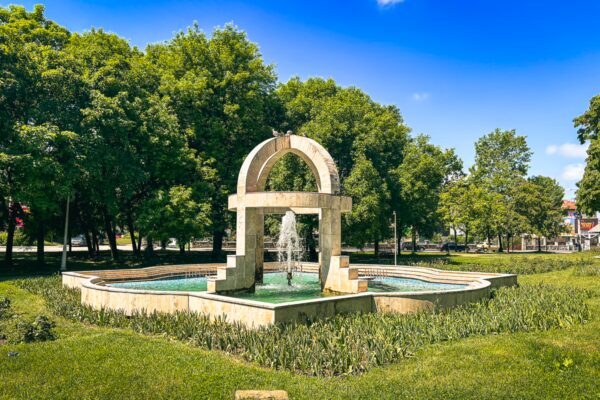 Fountain centerpiece surrounded by lush greenery at Mihai Bravu Garden in Craiova, Romania, under a clear blue sky.