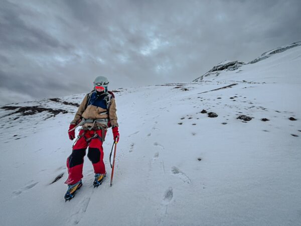 Climber in red and beige gear trekking through snow on Mount Cotopaxi, with dramatic gray skies overhead.