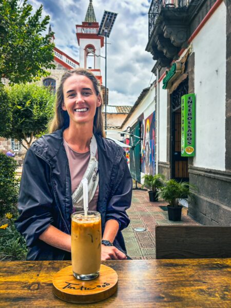 A woman smiles at a wooden table in front of a quaint café in Ibarra, Ecuador, with a refreshing drink and vibrant greenery around.
