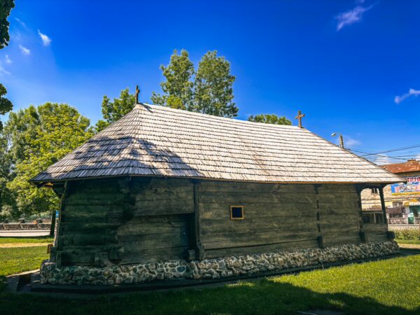 Rustic wooden structure with a thatched roof surrounded by greenery in Craiova, Romania, under a clear blue sky.