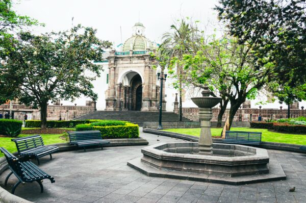 Plaza Grande in Quito features lush greenery, a central fountain, and benches, with a grand archway in the background.