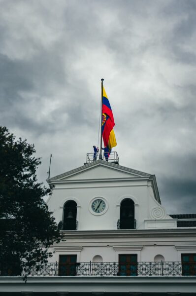 Carondelet Palace's white facade stands under a cloudy sky, featuring the vibrant Ecuadorian flag atop the building.