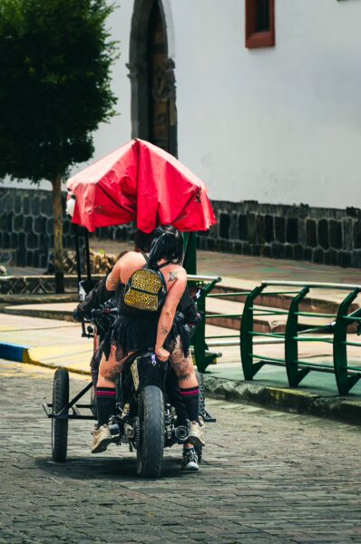 A person on a three-wheeled motorcycle rides past colorful street benches under a red umbrella in Ibarra, Ecuador.