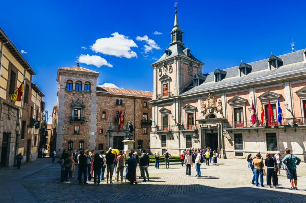 Crowd gathers in Plaza de la Villa, Madrid, showcasing stunning historic architecture and vibrant blue skies.