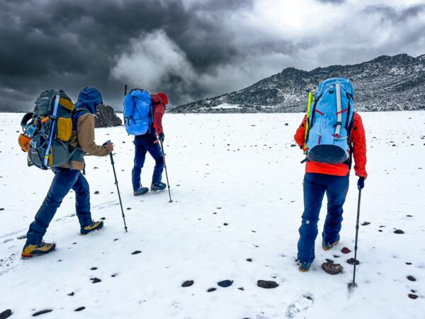 Hikers with colorful backpacks trek through snowy terrain near Chimborazo Volcano in Ecuador, under dynamic gray clouds.