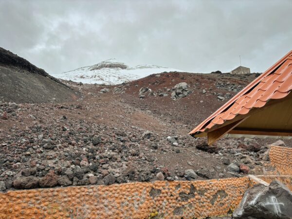 Snow-capped Mount Cotopaxi rises in the background, framed by rocky terrain and a rustic shelter at José Rivas Refuge in Ecuador.