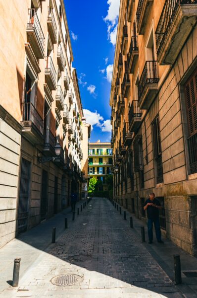 Sunlight illuminates a narrow street in Madrid, bordered by towering buildings with balconies and bright blue skies above.