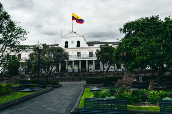 Colorful Ecuadorian flag flies over the white facade of a historic building, surrounded by greenery in Plaza Grande, Quito.