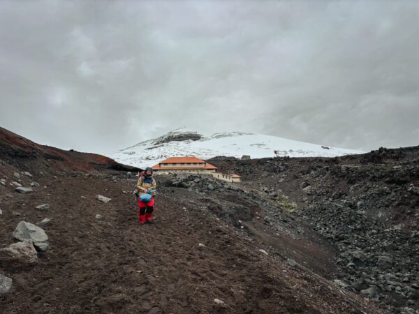 Hiker on a rocky path with snow-capped Mount Cotopaxi in the background, near the Jose Rivas Refuge in Ecuador.