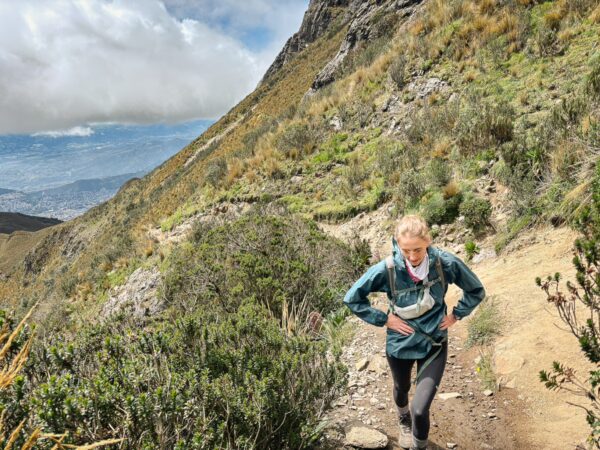 A hiker navigates a rugged trail surrounded by lush greenery on Pichincha Volcano, Ecuador, with scenic views in the distance.