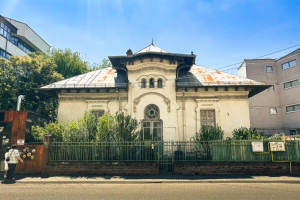 Charming white historic building with a distinctive roof, surrounded by greenery, set against a clear blue sky in Craiova, Romania.