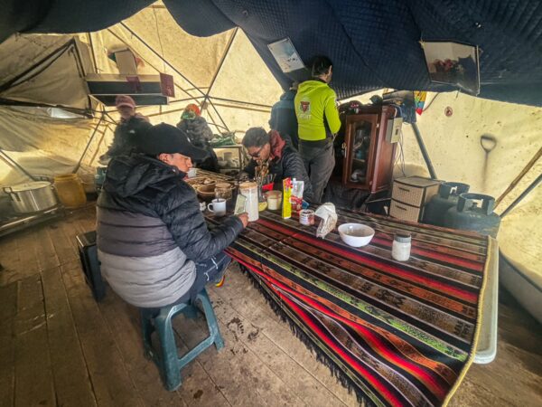 Travelers sit at a colorful table inside a high camp tent, enjoying warm drinks and food, surrounded by camping gear in Chimborazo, Ecuador.