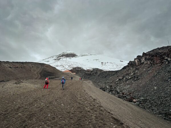 Hikers trek up the rugged trails of Mount Cotopaxi, with its snow-capped peak looming in the background under a cloudy sky.
