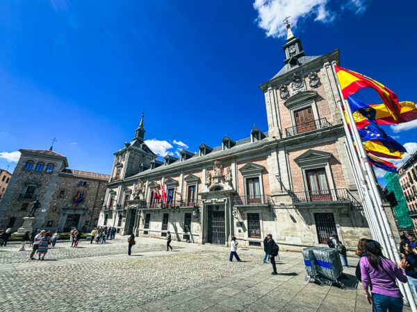 Historic architecture and vibrant flags in Plaza de la Villa, Madrid, Spain, under a bright blue sky.