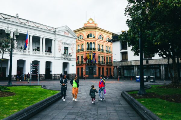 Colorful buildings frame Plaza Grande in Quito, Ecuador, with people strolling across the stone square under a cloudy sky.