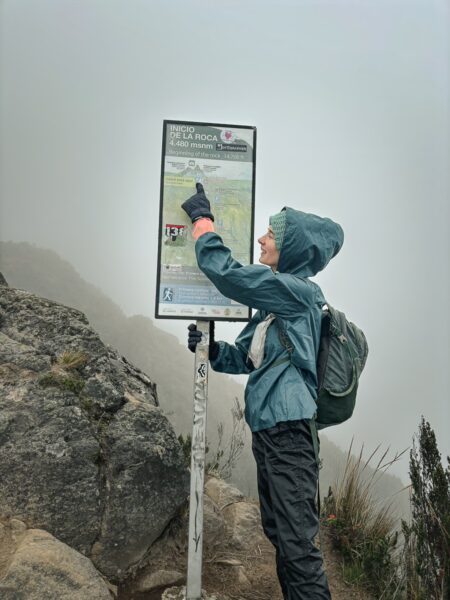 Hiker in a blue jacket examines a trail map beside rocky terrain, set against the misty backdrop of Imbabura Volcano, Ecuador.