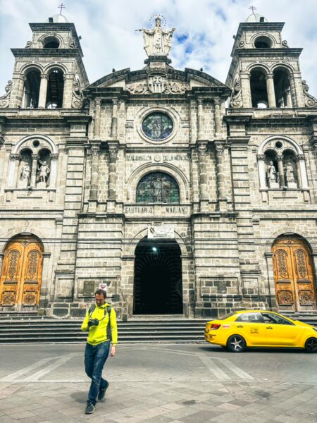La Merced Church stands prominently in Ibarra, Ecuador, showcasing intricate stonework and wooden doors, with a yellow taxi passing by.