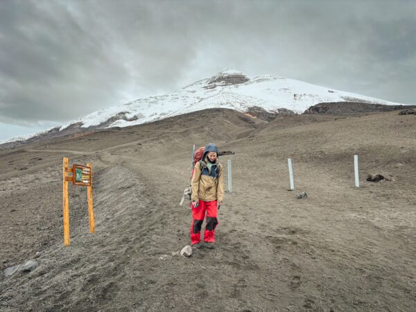 Hiker in bright red gear stands on a barren trail with Mount Cotopaxi looming in the background, covered in snow.