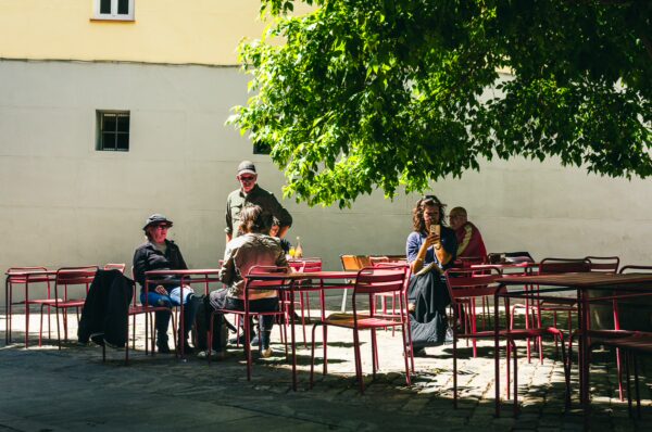 Relaxed atmosphere at Plaza del Biombo in Madrid, Spain, with patrons enjoying the shade of a large tree amid red chairs and tables.